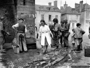 12 juin 1944 : Marie-Esther Duchenes, volontaire de la Croix Rouge avec des personnels d’une Engineer Special Brigade devant la fontaine de Sainte-Marie-du-Mont. Photo : US National Archives