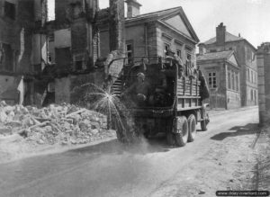 Deux sapeurs du 146th Engineer Combat Battalion, 1st (US) Army à bord d’un camion GMC, humidifient la route pour limiter les projections de poussière au passage des convois dans les ruines de Mortain. Photo : US National Archives