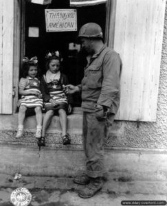 4 juillet 1944 : le sergent Walter P. Goworek offre des friandises à deux jeunes enfants du Molay-Littry habillées aux couleurs du drapeau américain. En arrière-plan sur la porte, un écritant "Vive les Américains" écrit en anglais avec une légère faute d'orthographe. Photo : US National Archives