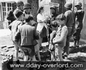 12 juin 1944 : Marie-Esther Duchenes, volontaire de la Croix Rouge avec des personnels d’une Engineer Special Brigade devant la fontaine de Sainte-Marie-du-Mont. Photo : US National Archives