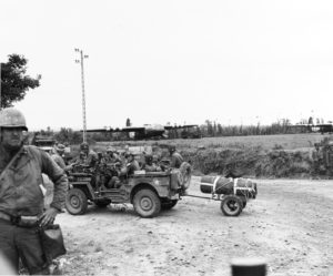 Peu après le Jour-J, des parachutistes de la 101st Airborne traversent un carrefour contrôlé par des MP à bord d'une Jeep. Des planeurs Waco sont visibles en arrière-plan. Derrière la Jeep se trouvent deux véhicules M29 Weasels et un camion GMC CCKW. Photo : US National Archives