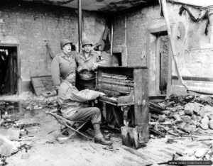 Des personnels civils américains du War Department portant l’uniforme marquent une pause autour d’un piano particulièrement abîmé par les bombardements à Valognes. Photo : US National Archives