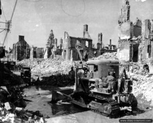 Un bulldozer dégage la rue de Poterie au milieu des ruines, le sol inondé suite à la rupture d’une canalisation d’eau à Isigny-sur-Mer. Photo : US National Archives