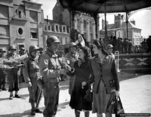 Sur la place de l'hôtel de ville de Cherbourg, deux normandes offrent des fleurs à un sous-officier américain membre de la fanfare. Photo : US National Archives