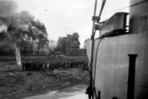 Des prisonniers allemands sont évacués sur la plage devant Bernières-sur-Mer tandis qu'une maison brûle en front de mer. Un bombardier allier survole la côte, de retour d'une mission avant de rejoindre l'Angleterre. Photo : Archives Canada