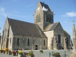 L'église Notre-Dame et le parvis de Sainte-Mère-Eglise (2010). Le mannequin du parachutiste John M. Steele est visible (dans les faits, il s'est accroché de l'autre côté du clocher le 6 juin 1944). Photo : Elliesram13