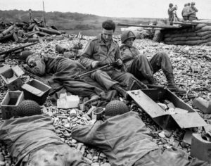 A Colleville-sur-Mer (Omaha Beach) en juin 1944, des sapeurs américains (dont l'un d'eux a camouflé l'arc de cercle blanc sur son casque) se reposent sur les galets de la plage, après avoir vidé des conserves de ration. Pendant qu'un des sapeurs nettoie le canon de son arme, des soldats en arrière-plan guident les navires de guerre lors des opérations de déchargement. Photo : US National Archives