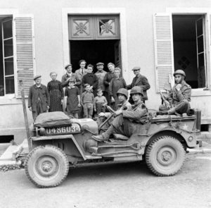 Dans Cerisy-la-Forêt, devant le n°6 rue Robert Le Magnifique, les habitants posent derrière une Jeep transportant des militaires de la 2nd (US) Infantry Division et un soldat français. A noter : la barre métallique coupe-câble à l'avant du véhicule. Photo : US National Archives