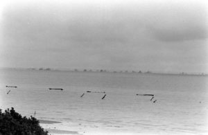 Au large de Grandcamp-les-Bains, des navires de guerre alliés croisent en large. Sur la plage, des défenses de plage baptisées "Holzfähle" et surmontées de mines antichar "Tellermine". Photo : US National Archives