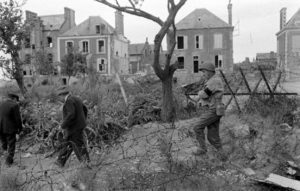 Rue des Ecoles à Grandcamp-les-Bains, le sergent Roland des Autels (à droite) du CIC (Counter Intelligence Corps) s'avance dans le périmètre défensif du port. Avec la canne, au milieu, Pierre Ferrari, conseiller municipal de Grandcamp-les-Bains. Photo : US National Archives