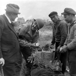 Le sergent Roland des Autels du CIC (Counter Intelligence Corps) et Pierre Ferrari (à gauche), conseiller municipal de Grandcamp, observent avec deux civils (à droite) le contenu de musettes abandonnées par les Allemands sur une position défensive des abords du port, à l'emplacement de l'actuel chemin de la Villa Mathieu. Une mitrailleuse française Reibel MAC 31, utilisée par les Allemands, est visible en arrière plan avec ses chargeurs "camemberts". Photo : US National Archives