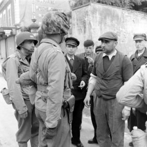 Un officier supérieur américain, avec un casque recouvert de camouflage, échange avec des habitants de Grandcamp-les-Bains, sur l'actuelle rue de la Libération. Au centre, habillé de noir, se trouve monsieur Le Galou, du syndic des gens de mer. A noter : l'officier porte du pain sous son bras.