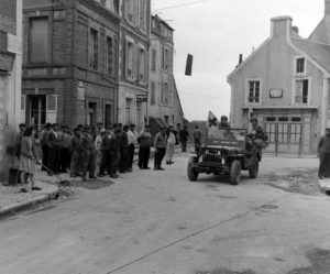 Une Jeep médicalisée transporte un blessé sur l'actuelle rue Aristide Briand à Grandcamp après la libération, sous les yeux des habitants rassemblés. La rue de la Marine est bloquée par un mur antichar. Photo : US National Archives