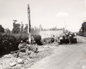 Des parachutistes américains de la 101st (US) Airborne Division interdisent le carrefour de La Fourchette, à proximité de Carentan, avec un canon antichar de 57 mm. En arrière-plan, un canon d'assaut Stug IV du SS-Panzer-Abteilung 17. Photo : US National Archives