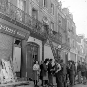 Place de la République à Carentan, des civils installent des drapeaux aux balcons des habitations. Deux parachutistes de la 101st (US) Airborne Division sont au milieu d'eux. Photo : US National Archives