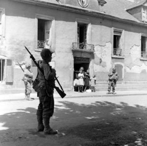 Des soldats aérotransportés américains patrouillent le long de la rue du quai à vin à Carentan devant les habitants. Ils portent la baïonnette au canon, signe que du combat rapproché peut survenir à tout moment. Photo : US National Archives