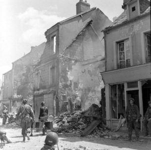 Des parachutistes américains de la 101 (US) Airborne Division dans la rue Holgate à Carentan, au pied des bâtiments détruits par les bombardements. Des civils quittent les lieux, accompagnés de gendarmes français. Photo : US National Archives