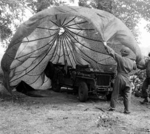 Juin 1944 : des parachutistes américains camouflent une Jeep à l'aide d'un parachute bariolé, au campement provisoire de l’état-major de la 101st (US) Airborne Division, situé au bassin à flot à Carentan. Photo : US National Archives