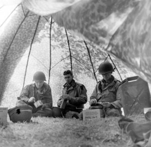 Juin 1944 : des parachutistes américains se restaurent à l'ombre d'un parachute, au campement provisoire de l’état-major de la 101st (US) Airborne Division, situé au bassin à flot de Carentan. Photo : US National Archives