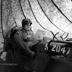 Juin 1944 : un parachutiste américain lit un journal à bord d'une Jeep camouflée par un parachute, au campement provisoire de l’état-major de la 101st (US) Airborne Division, situé au bassin à flot à Carentan. Photo : US National Archives