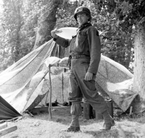 Juin 1944 : un parachutiste américain (à hauteur des abris aménagés avec des parachutes), transporte un petit réservoir souple d'eau, au campement provisoire de l’état-major de la 101st (US) Airborne Division, situé au bassin à flot à Carentan. Photo : US National Archives