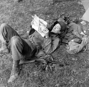 Juin 1944 : un parachutiste américain se distrait en lisant l'édition de Juin 1944 du magazine Sparkler Comics, son pistolet-mitrailleur Thompson posé à côté de lui, au campement provisoire de l’état-major de la 101st (US) Airborne Division, situé au bassin à flot à Carentan. Photo : US National Archives