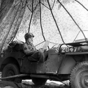 Juin 1944 : un parachutiste américain lit un journal à bord d'une Jeep camouflée par un parachute, au campement provisoire de l’état-major de la 101st (US) Airborne Division, situé au bassin à flot à Carentan. Photo : US National Archives