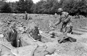 12 juillet 1944 : des prisonniers allemands creusent des tombes pour leurs camarades tués durant les combats, sous le contrôle de soldats américains au cimetière militaire d'Orglandes. Photo : US National Archives
