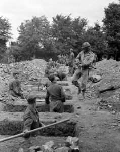 12 juillet 1944 : des prisonniers allemands creusent des tombes pour leurs camarades tués durant les combats, sous le contrôle de soldats américains au cimetière militaire d'Orglandes. Photo : US National Archives