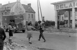 Juillet 1944 : deux habitantes de la ville de Caen (dont Denise Costil, avec un panier) offrent des cerises aux libérateurs canadiens, rue du gérénal Moulin. Un camion Fordson 7V circule à proximité. A droite de la photo, la station service "Relais de Caen". Photo : George Rodger pour LIFE Magazine