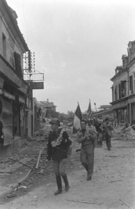 Rue de la gare à Caen en juillet 1944, des résistants français appartenant à la compagnie Scamaroni. En tête de la colonne : Léonard Gille, suivi par Serge Goguel. Ils portent les drapeaux français et britannique notamment pour éviter un tir fratricide de la part des Alliés. Photo : George Rodger pour LIFE Magazine