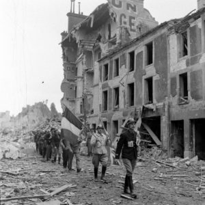 Rue de la gare à Caen en juillet 1944, des résistants français appartenant à la compagnie Scamaroni. En tête de la colonne : Léonard Gille, suivi par René Duchez. Ils portent les drapeaux français et britannique notamment pour éviter un tir fratricide de la part des Alliés. Photo : George Rodger pour LIFE Magazine