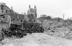 A Caen, des motocyclistes et pilotes stationnent avec leurs véhicules dans les ruines de la rue d'Haleine (devenue l'Esplanade de la Paix), au pied du château, en juillet 1944. Ils appartiennent à la colonne chargée du transport et de l'escorte du Lieutenant General John T. Crocker, commandant le 1st Corps. Photo : George Rodger pour LIFE Magazine