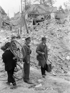 Dans les décombres de Caen en juillet 1944, le Lieutenant General John T. Crocker, commandant le 1st Corps, inspecte les lieux en compagnie de civils. A noter : la carcasse d'un véhicule à l'étage du bâtiment détruit en arrière-plan. Photo : George Rodger pour LIFE Magazine