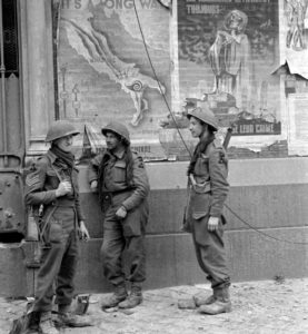 Juillet 1944 : trois soldats britanniques de la 3rd (GB) Infantry Division à hauteur du marché couvert de Caen. Sur le mur, des affiches de propagande allemandes : à gauche, l'Axe se moque de la vitesse de progression des Alliés en Italie, qu'ils comparent à un escargot ("It's a long way to Rome" : "qu'il est long le chemin jusqu'à Rome"). A droite, l'affiche dénonce les bombardements alliés avec la référence à Jeanne d'Arc, brûlée à Rouen, après les raids alliés qui ont détruit la ville du 30 mai au 5 juin 1944. Photo : George Rodger pour LIFE Magazine.
