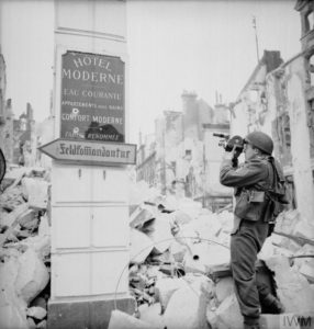 Le sergent J. H. Goddard, cameraman britannique,, filme les vestiges de l'hôtel Moderne avec sa caméra Eyemo. L'hôtel était situé boulevard Saint-Pierre (aujourd'hui boulevard des Alliés) à Caen. Un panneau de signalisation allemand indique la direction de la Feldkommandantur 723 de Caen, situé dans l'hôtel Malherbe de la place Foch. Photo : IWM B 6804