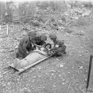 10 juillet 1944 : un blessé anglais de la 3rd (GB) Infantry Division, installé dans un brancard dans le secteur de Caen, est réconforté par ses camarades avant d'être évacué vers un centre de triage. Photo : IWM B 6829