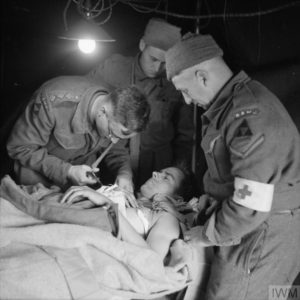 10 juillet 1944 : un blessé anglais de la 3rd (GB) Infantry Division est ausculté par un capitaine médecin du Royal Army Medical Corps, accompagné par deux secouristes, dans un centre de triage du secteur de Caen. Photo : IWM B 6834