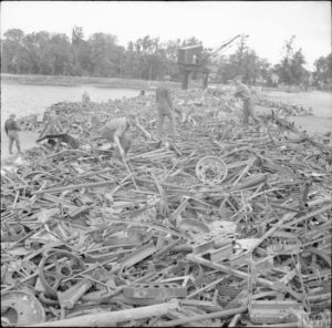 Juillet 1944 : sur les quais des chantiers navals de Caen, des prisonniers allemands empilent des vestiges d'armements, équipements et véhicules de guerre qui seront ensuite transportés par bateau jusqu'en Angleterre. Photo : IWM BU 7881