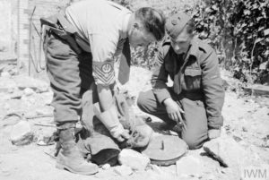 A Caen, les militaires du No. 6237 Bomb Disposal Flight appuient le nettoyage des décombres en neutralisant les pièges explosifs mis en place par les Allemands. Le Flight Sergeant D. Simons Didcot (à gauche) explique à un membre des forces aériennes britanniques le mode de fonctionnement d'un piège composé d'une mine antichar "Tellermine" et d'un manteau. Les deux éléments étaient reliées et le manteau recouvrait la mine. Photo : IWM CL 748