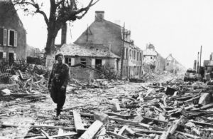 Juillet 1944 : le Sergeant Jimmy Mapham, photographe au sein de la 5th Army Film & Photographic Unit (AFPU), traverse les décombres de la périphérie de Caen, son appareil photo autour du cou. Photo : IWM HU 109817