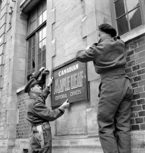 11 juillet 1944 : rue Desmolombe à Caen, le Lieutenant-Colonel R.S. Malone (avec le pistolet à la ceinture) accroche l’enseigne du journal canadien "Maple Leaf" sur un mur du bâtiment de "La Presse Caennaise". Il est accompagné du Major Mac Farlane, rédacteur en chef, et du Major Seth Halton. Photo : Library and Archives Canada