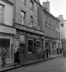 Des militaires américains et britanniques ainsi que des civils devant la devanture du magasin "Tout pour la Photo" de L. Leprunier situé au numéro 53 de la rue Saint-Malo à Bayeux, durant l'été 1944 après la libération. Photo : LIFE Magazine