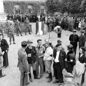 Place aux Pommes à Bayeux le 14 juillet 1944, avant la cérémonie en hommage aux Morts. De gauche à droite au premier plan : Léonard Gille (casque), Serge Goguel (souriant), René Duchez (casqué avec le brassard tricolore), Georges Poilane (bras plâtré) et Louise Boitard, résistants de la compagnie Scamaroni. Des Français du 1er bataillon de fusiliers marins commandos montent la garde autour du monument aux Morts, aux ordres du lieutenant de vaisseau Alexandre Lofi (avec son pistolet automatique à la ceinture). Photo : George Rodger pour LIFE Magazine