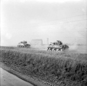 Deux chars Sherman appartenant à la 11th Armoured Division contournent Ranville par l'est le 18 juillet 1944 pendant l'opération Goodwood. Les ruines de l'église de l'Assomption Notre-Dame sont visibles en arrière-plan. Photo : IWM