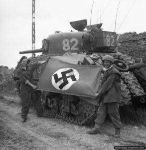 10 juillet 1944 : l’équipage d’un char Sherman présente un drapeau nazi pris pendant l’attaque de Caen (le Lance Corporal S. James et le Sergeant H. Coe de la 27th Armoured Brigade. Photo : IWM