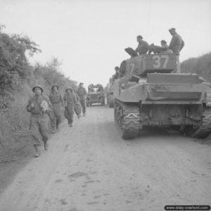 8 juillet 1944 : des soldats anglais marchent à proximité d’un char Sherman Firefly VC appartenant au 144th Regiment Royal Armoured Corps de la 33rd Armoured Brigade dans le secteur du bois de Lébisey à proximité de Hérouville-Saint-Clair pendant l’opération Charnwood. Photo : IWM