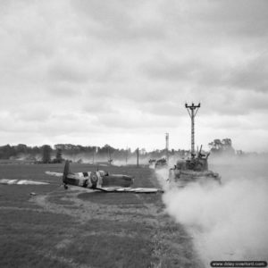 17 juin 1944 : des chars Sherman progressent dans un champ à proximité d’un chasseur Spitfire ayant fait un atterrissage d’urgence près de Tilly-sur-Seulles. Photo : IWM
