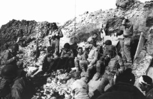 Des Rangers américains du 2nd Ranger Battalion savourent un moment de répit après de durs combats livrés à la Pointe du Hoc. Deux parachutistes du I/506th Parachute Infantry Regiment (101st Airborne Division) ont participé aux combats : Ray Crouch et Len Goodgall. Parachutés par erreur dans le secteur, ils ont renforcé les Rangers avant de rejoindre leur unité après le 8 juin 1944. Len Goodgall et son insigne de division sont visibles sur cette photo, sous le drapeau avec la croix rouge. Un commando anglais, l'officier de liaison Thomas Trevor, peut également être vu en bas à droite, avec un bandeau blanc sur le visage. Photo : US National Archives