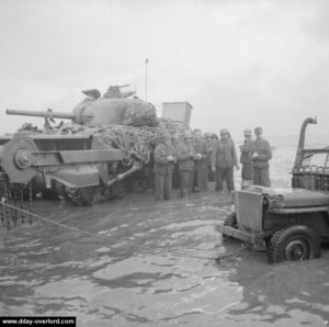 Des prisonniers allemands attendent l'ordre d'évacuation les pieds dans l'eau et à l'abri d'un char Sherman Crab Flail abandonné, tandis qu'une Jeep est remorquée. Photo : IWM